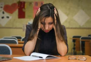 frustrated teacher sitting at wood desk or table in classroom holding her head in her hands and looking at a notebook used for header on blog post on assessments in secondary reading intervention