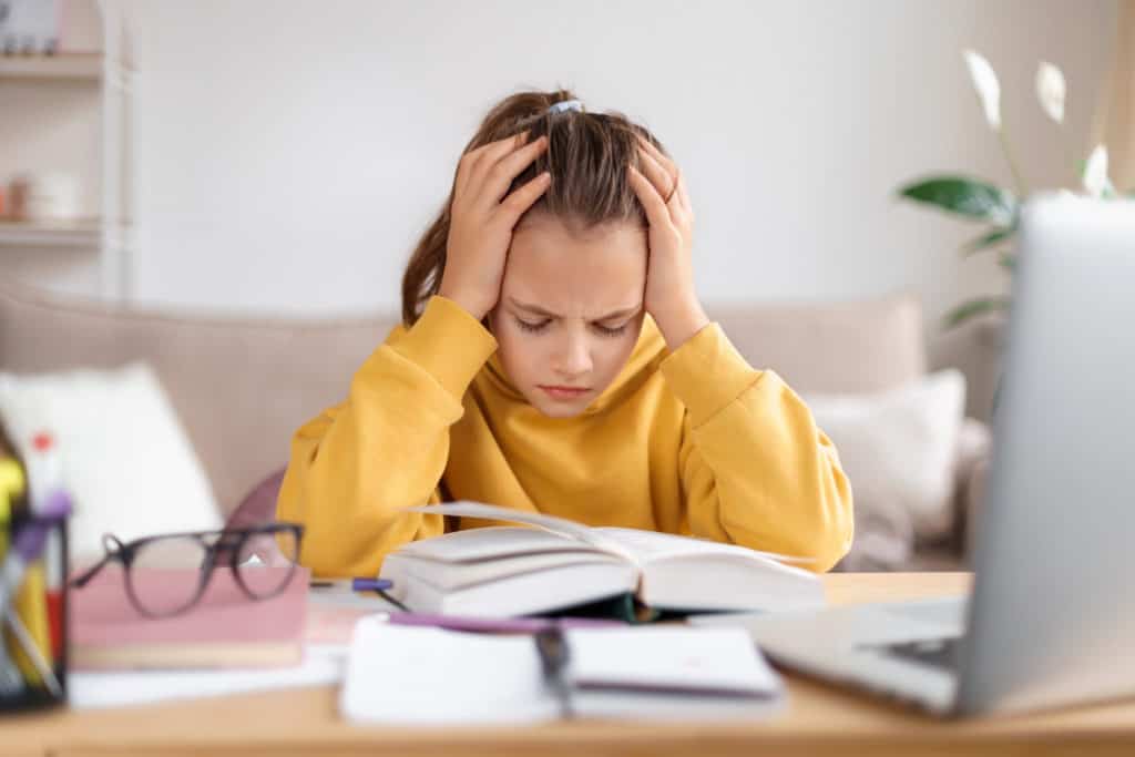 struggling reader girl in yellow hands on head seated in front of books looking defeated
