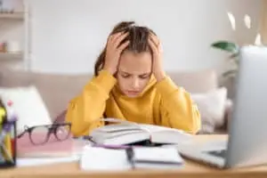 struggling reader girl in yellow hands on head seated in front of books looking defeated